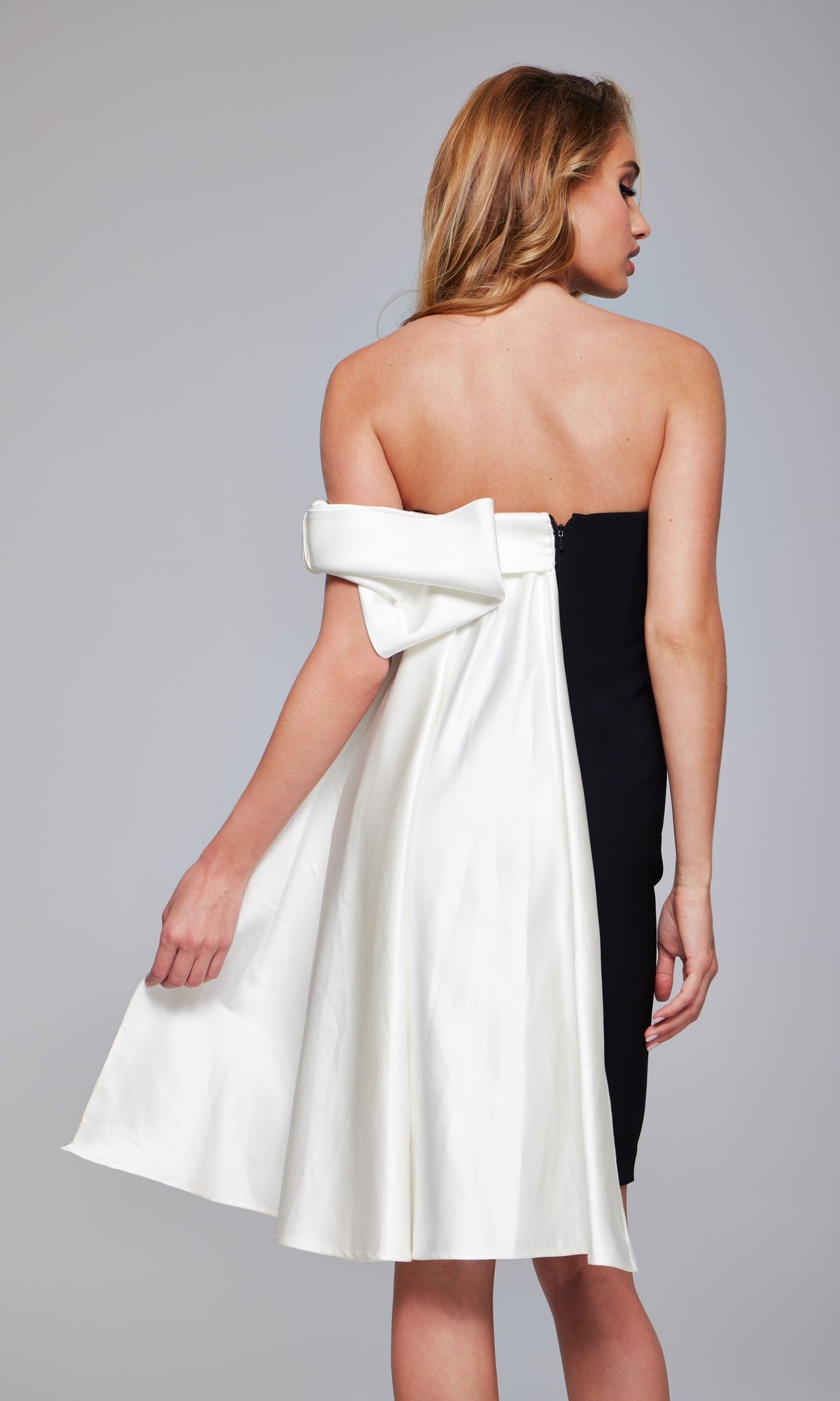 Woman in a Jovani Draped Off-Shoulder Short Hoco Dress 37392, featuring a strapless black design with a large white bow detail on the back, stands facing away from the camera against a plain background.